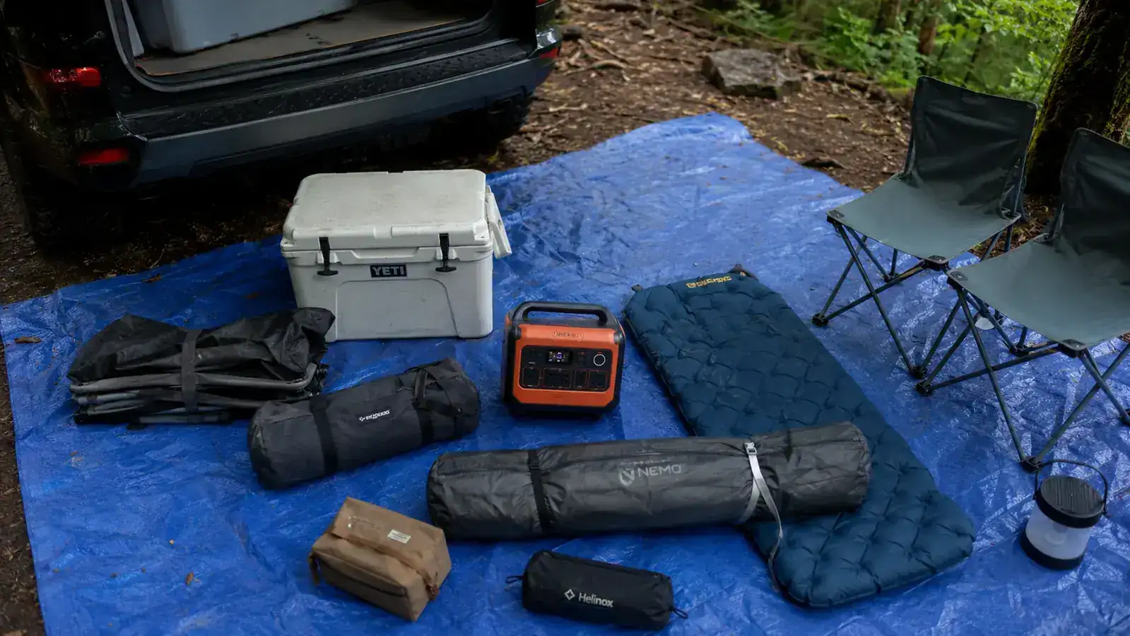 Overhead view of camping gear including a portable power station laid out on a tarp beside an open SUV hatch