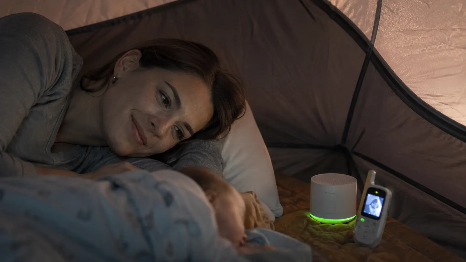 Close-up of a white-noise machine and baby monitor side by side on a tent floor, green charging indicator light glowing