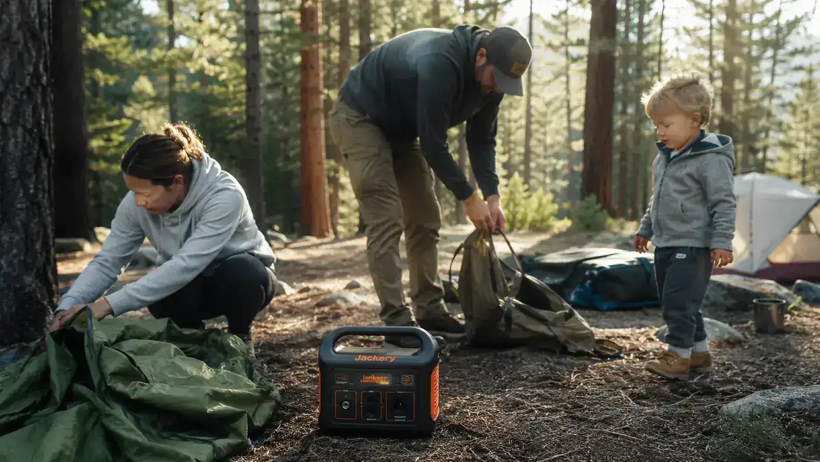 Family packing up a campsite in the morning, Jackery power station visible next to a folded tent, toddler watching nearby