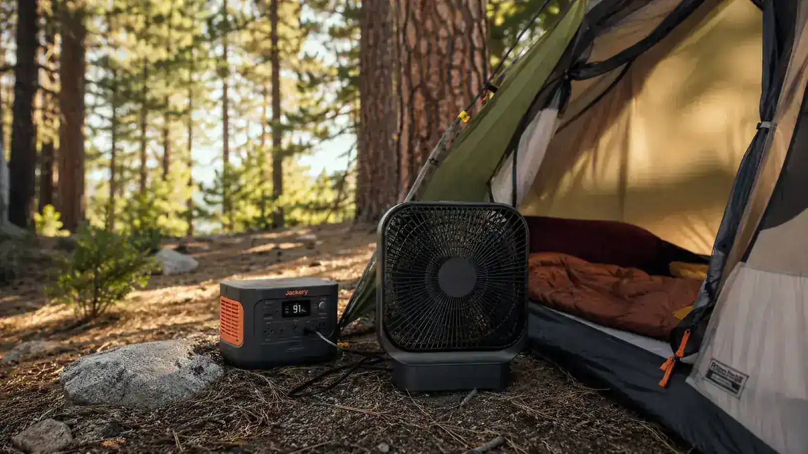Jackery Explorer 300 sitting next to a tent at a campsite with a small box fan plugged in, morning light through pine trees