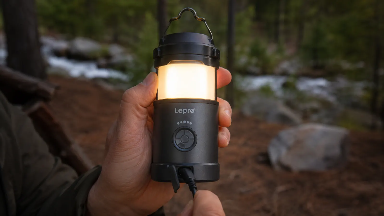 Hand holding a compact rechargeable LED camping lantern, USB-C cable plugged into the side, lantern lit in warm mode